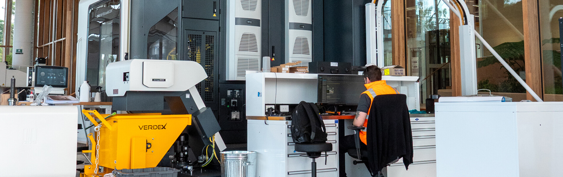 A man in a high-vis vest works on a computer surrounded by machinery.