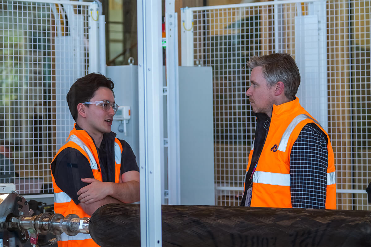 Two men wearing high-vis vests talking and standing next to a machine.